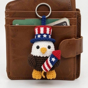May include: A crocheted keychain featuring a bald eagle wearing a red, white, and blue top hat with stars. The eagle has a yellow beak and feet, a white head, and brown body, and is holding a red, white, and blue firework with a white star.