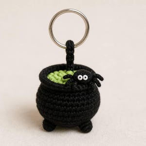 May include: A black crocheted cauldron keychain featuring a bright green textured interior and a small black spider with two white eyes attached to the rim. The cauldron has three small feet and is connected to a silver metal split ring.