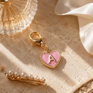 Pode incluir: Um berloque de metal em tom dourado com um pingente em forma de coração com um centro de esmalte rosa e a letra A em ouro. O berloque está preso a um fecho lagosta dourado, projetado para ser facilmente fixado em vários itens, como bolsas ou joias.