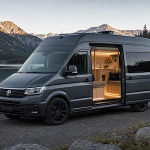Puede incluir: Una furgoneta camper Volkswagen de color gris oscuro con la puerta lateral corredera abierta, revelando un interior de madera clara con armarios integrados, asientos e iluminación cálida. El vehículo tiene ruedas negras y está aparcado en un camino.