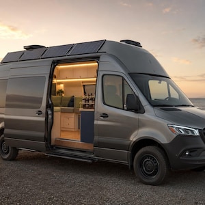 Op de afbeelding: Een grijze Mercedes-Benz Sprinter camper met zonnepanelen op het dak en een open schuifdeur aan de zijkant die een verlicht interieur onthult met lichtgekleurde kasten, groene zitplaatsen en een kleine keuken bij de oceaan.