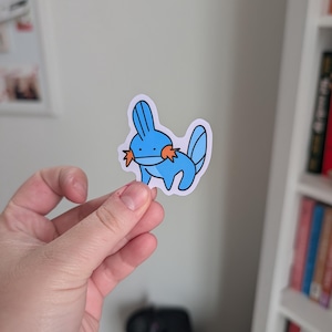 May include: A die-cut vinyl sticker featuring a cartoon Mudkip character. The creature is light blue with a darker blue fin on its head and tail, and orange star-shaped cheek gills. It has a simple black outline and a white border.