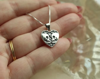 May include: A silver necklace with a heart-shaped pendant featuring two skulls facing each other. The pendant is decorated with small stars and crescent moons.