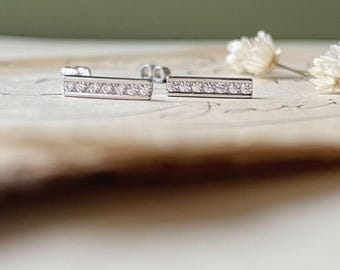 May include: Silver bar stud earrings, each featuring a line of small, clear gemstones. The earrings are presented on a light surface, with a soft, blurred background. The studs have a simple, elegant design, suitable for everyday wear.