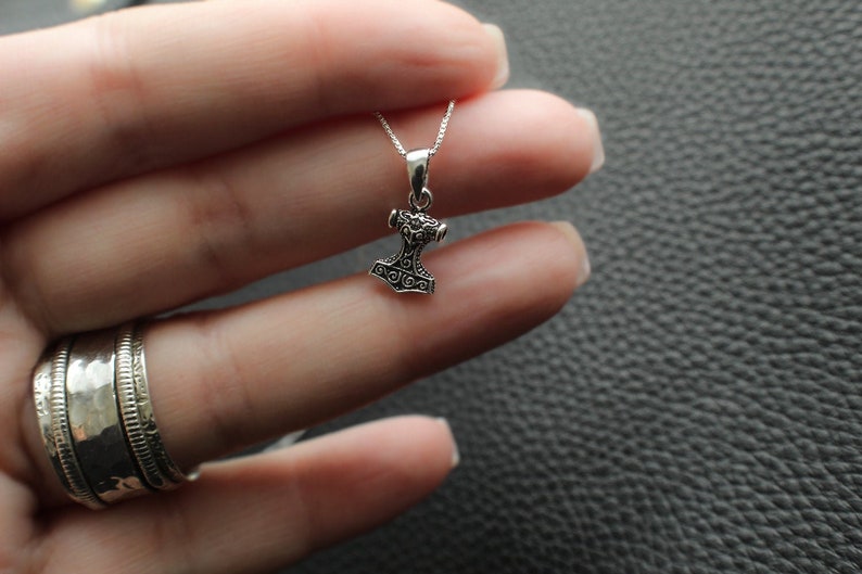 May include: A silver pendant necklace featuring a detailed Mjolnir hammer charm. The hammer is a symbol of Thor, the Norse god of thunder.