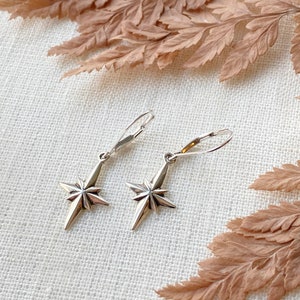 May include: A pair of silver star-shaped dangle earrings with a simple hook closure. The earrings are on a white fabric background.