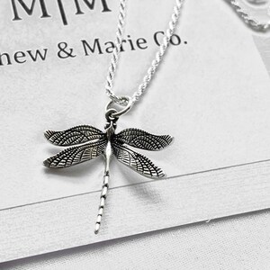 May include: A silver chain necklace with a detailed dragonfly pendant. The pendant is silver and has a textured design. The necklace is on a white background with the text "Mathew & Marie Co."