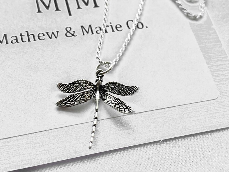 May include: A silver chain necklace with a detailed dragonfly pendant. The pendant is silver and has a textured design. The necklace is on a white background with the text "Mathew & Marie Co."