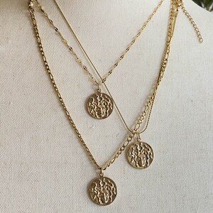 Peut inclure: Trois colliers en cha&icirc;ne dor&eacute;e avec des m&eacute;daillons dor&eacute;s repr&eacute;sentant un visage de femme en relief. Les m&eacute;daillons sont de tailles diff&eacute;rentes et sont superpos&eacute;s.