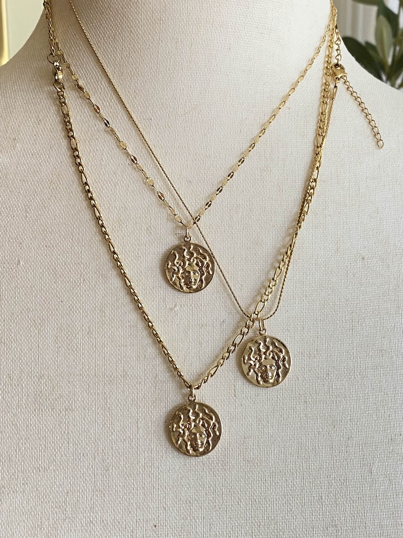 Peut inclure: Trois colliers en cha&icirc;ne dor&eacute;e avec des m&eacute;daillons dor&eacute;s repr&eacute;sentant un visage de femme en relief. Les m&eacute;daillons sont de tailles diff&eacute;rentes et sont superpos&eacute;s.