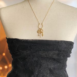 May include: A gold-colored necklace with a horseshoe pendant and various charms, including a heart, star, and pearl. The necklace is displayed on a mannequin wearing a black lace top.