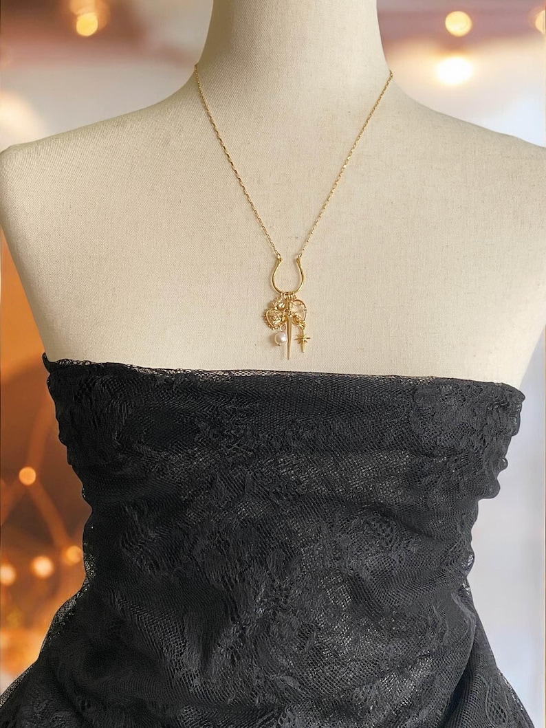 May include: A gold-colored necklace with a horseshoe pendant and various charms, including a heart, star, and pearl. The necklace is displayed on a mannequin wearing a black lace top.
