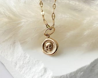 May include: A gold-colored necklace featuring a pendant with a skull design. The pendant is round and has a detailed skull image in the center. The chain is delicate and made of small, connected links. The necklace is displayed on a white surface.