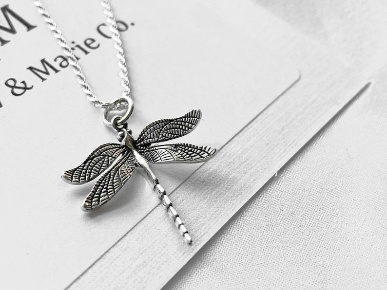 May include: Silver necklace with a detailed dragonfly pendant. The necklace is on a white background with the text "M & Marie Co."
