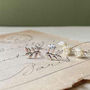 May include: Silver leaf stud earrings with a delicate design. The earrings are displayed on a vintage paper with script and dried white flowers. The earrings have a polished finish and are a nature-inspired jewelry piece.