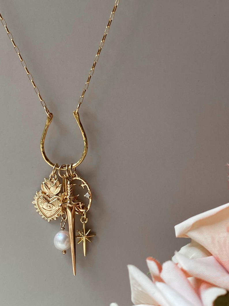 May include: Gold-toned necklace with a horseshoe-shaped pendant. The pendant features charms including a heart, sword, star, and a pearl. The necklace is displayed against a neutral background with a hint of pink flowers.
