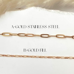 May include: Two gold-colored chain necklaces are displayed against a white background. The top chain is labeled "A-GOLD STAINLESS STEEL" and features large oval links. The bottom chain, labeled "B-GOLD FILL", has smaller rectangular links. The chains are presented in a jewelry product shot.