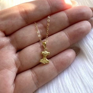 May include: A gold chain necklace with a small gold pendant shaped like a hammer. The pendant has intricate details and a textured surface.