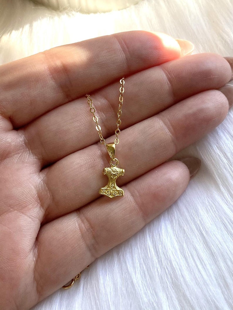 May include: A gold chain necklace with a small gold pendant shaped like a hammer. The pendant has intricate details and a textured surface.