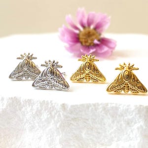 May include: Four moth-shaped stud earrings in silver and gold tones are displayed on a white surface. The earrings have detailed wing and body designs. A pink flower is in the background, adding a touch of color.
