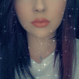 May include: A close-up of a person's face with dark hair and a silver necklace with a small pendant. The person is wearing a white shirt and has a soft, natural makeup look.