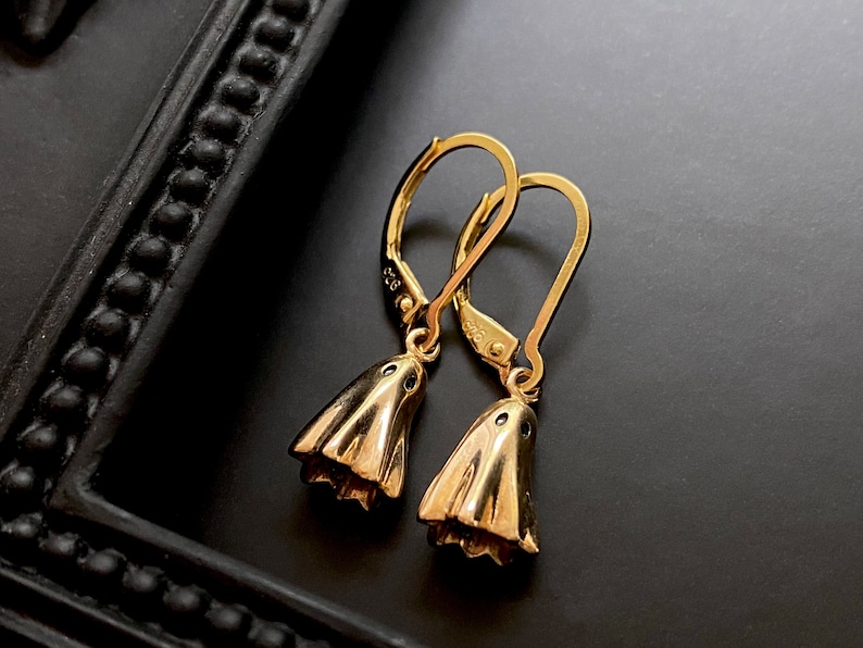 May include: A pair of gold dangle earrings with a bell-shaped design. The earrings have a simple, elegant style.