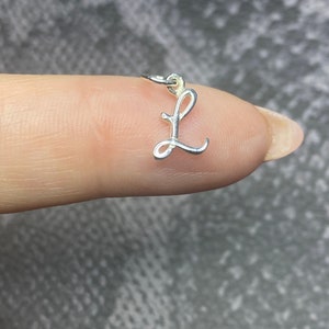 May include: A small, silver-coloured charm in the shape of the letter "L". The charm is crafted in a cursive font and has a small loop at the top for attachment. The charm is shown against a blurred background.