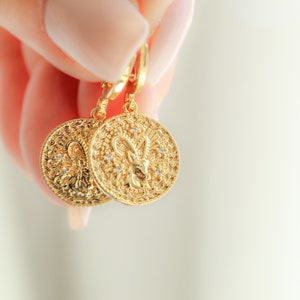 May include: Gold-tone earrings with a detailed design featuring a zodiac symbol. The earrings are hanging from a hook and are held by a hand.