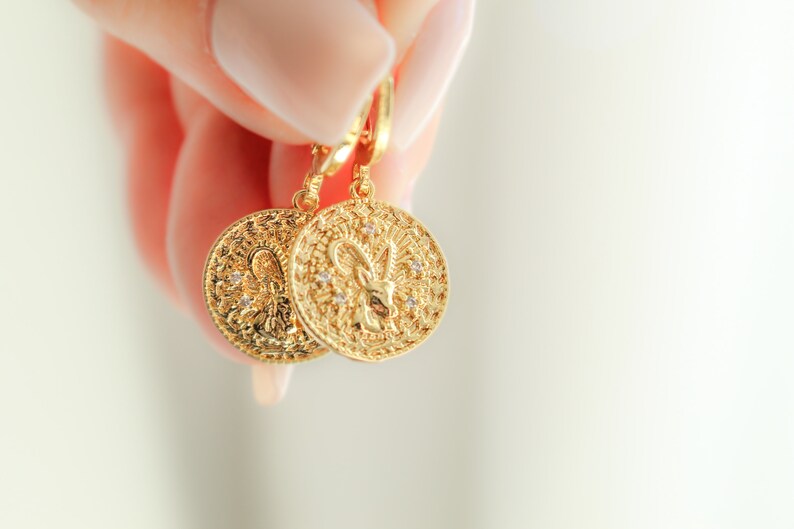May include: Gold-tone earrings with a detailed design featuring a zodiac symbol. The earrings are hanging from a hook and are held by a hand.
