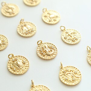 May include: Twelve gold zodiac charms with a white background. Each charm features a different zodiac symbol with small white stones.