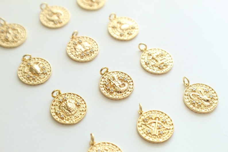 May include: Twelve gold zodiac charms with a white background. Each charm features a different zodiac symbol with small white stones.