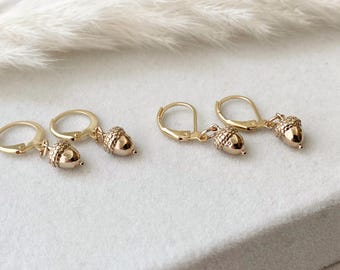 May include: Gold-colored acorn earrings with lever-back closures. The earrings feature detailed acorn pendants, showcasing a textured cap and a smooth body. The earrings are displayed on a light-colored surface, with soft, feathery elements in the background.