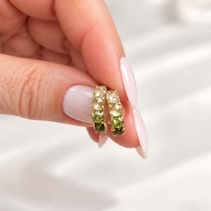 May include: A pair of small gold hoop earrings featuring a gradient of round faceted gemstones. The stones transition in color from light yellow and pale green at the top to deep olive and dark forest green at the bottom of each circular hoop.