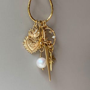 May include: Gold-toned charm necklace featuring a variety of pendants. Charms include a heart with an eye, a sword, a pearl, a star, and a crescent moon with a small gem. The necklace is suspended from a textured gold hoop.