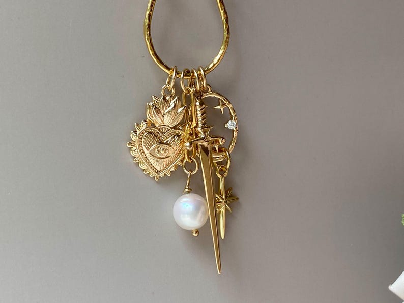 May include: Gold-toned charm necklace featuring a variety of pendants. Charms include a heart with an eye, a sword, a pearl, a star, and a crescent moon with a small gem. The necklace is suspended from a textured gold hoop.