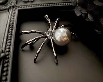 May include: A silver spider brooch featuring a large, grey pearl body and black detailing. The spider's legs have a metallic finish. The brooch is set against a dark backdrop within a decorative frame, evoking a gothic or Halloween aesthetic.