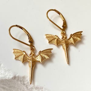 May include: A pair of gold-colored dragon earrings. Each earring features a detailed dragon pendant with outstretched wings and a long, pointed tail. The earrings have lever-back closures and are displayed on a white surface.