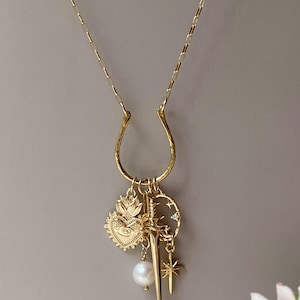 May include: Gold-toned necklace with a horseshoe-shaped pendant and multiple charms. Charms include a heart with an eye, a sword, a pearl, a star, and a crescent moon with stars. The necklace has a delicate chain.