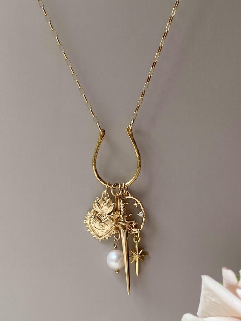 May include: Gold-toned necklace with a horseshoe-shaped pendant and multiple charms. Charms include a heart with an eye, a sword, a pearl, a star, and a crescent moon with stars. The necklace has a delicate chain.