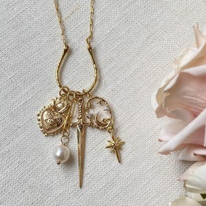 May include: Gold-toned pendant necklace featuring a horseshoe-shaped bail. The charms include a heart, a sword, a pearl, a crescent moon, and a star. The necklace is displayed on a white linen surface, with a soft pink rose in the background.
