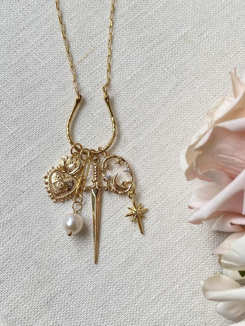 May include: Gold-toned pendant necklace featuring a horseshoe-shaped bail. The charms include a heart, a sword, a pearl, a crescent moon, and a star. The necklace is displayed on a white linen surface, with a soft pink rose in the background.