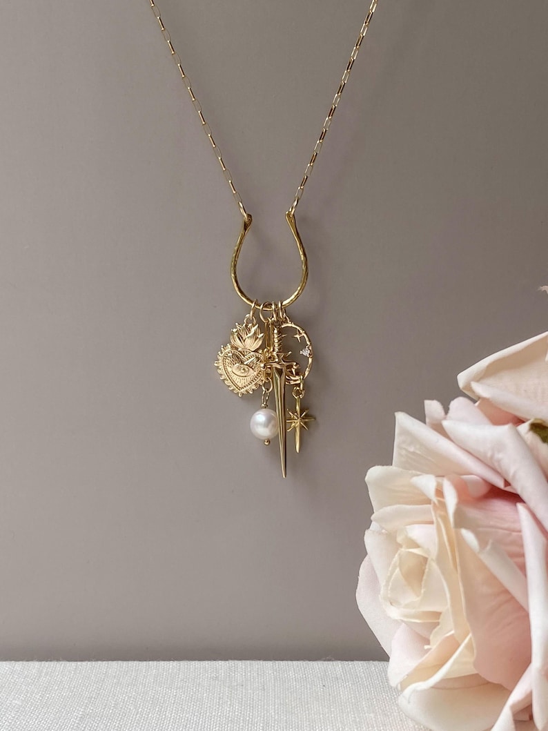 May include: A gold-colored necklace with a horseshoe-shaped pendant and multiple charms. Charms include a heart, a sword, a star, and a pearl. The necklace is displayed against a gray background with a soft pink rose in the corner.