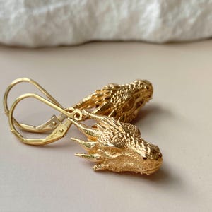 May include: A pair of gold-colored dragon head earrings. The earrings feature detailed dragon heads with textured scales and pointed features. The lever-back closures are also gold-colored. The earrings are displayed on a neutral background.