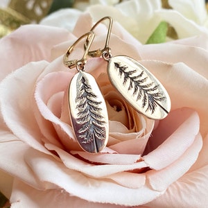 May include: A pair of gold earrings with a delicate design of a pine tree on each earring. The earrings are oval shaped and have a simple hook closure.