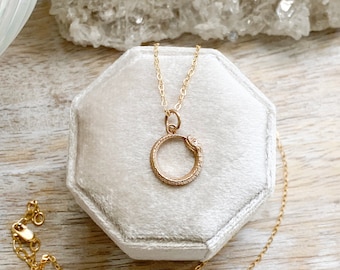 May include: Gold snake chain necklace with a small, gold snake charm in the shape of a circle. The necklace is displayed in a white velvet box.