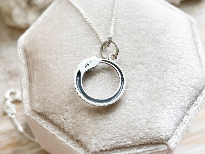 May include: A silver snake necklace with a circular pendant. The pendant has a black enamel finish and the words "925 N" are engraved on the back.