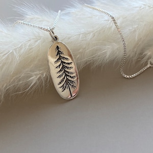 May include: A silver-toned necklace featuring an oval pendant with an etched pine tree design. The pendant hangs from a delicate chain. The background is a soft, blurred white, creating a minimalist aesthetic. The necklace is a piece of jewelry.