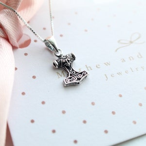 May include: A silver necklace with a small, detailed Mjolnir pendant. The pendant is a replica of Thor's hammer from Norse mythology. The necklace is on a white background with pink polka dots. The text "Matthew and Mary Jewelry Atelier" is visible in the background.