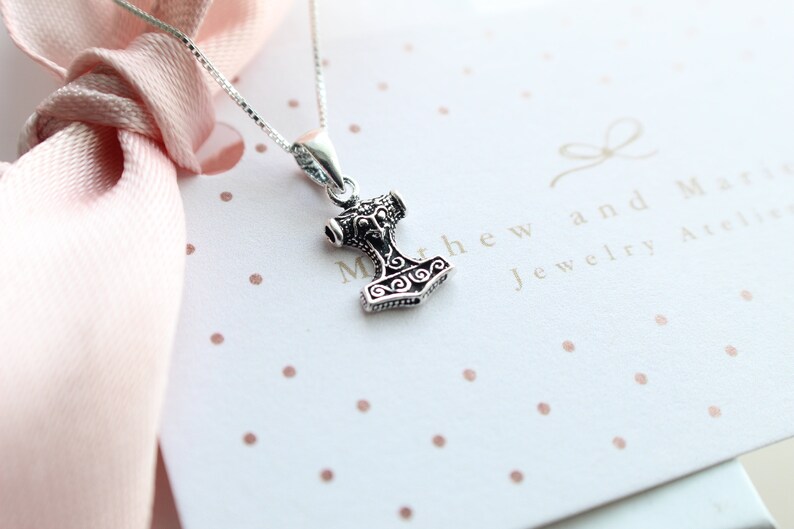 May include: A silver necklace with a small, detailed Mjolnir pendant. The pendant is a replica of Thor's hammer from Norse mythology. The necklace is on a white background with pink polka dots. The text "Matthew and Mary Jewelry Atelier" is visible in the background.