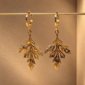 May include: A pair of gold-toned hoop earrings featuring dangling leaf-shaped charms. Each earring consists of a polished circular hoop connected to a detailed, multi-lobed leaf pendant with a reflective metallic finish and defined veins.